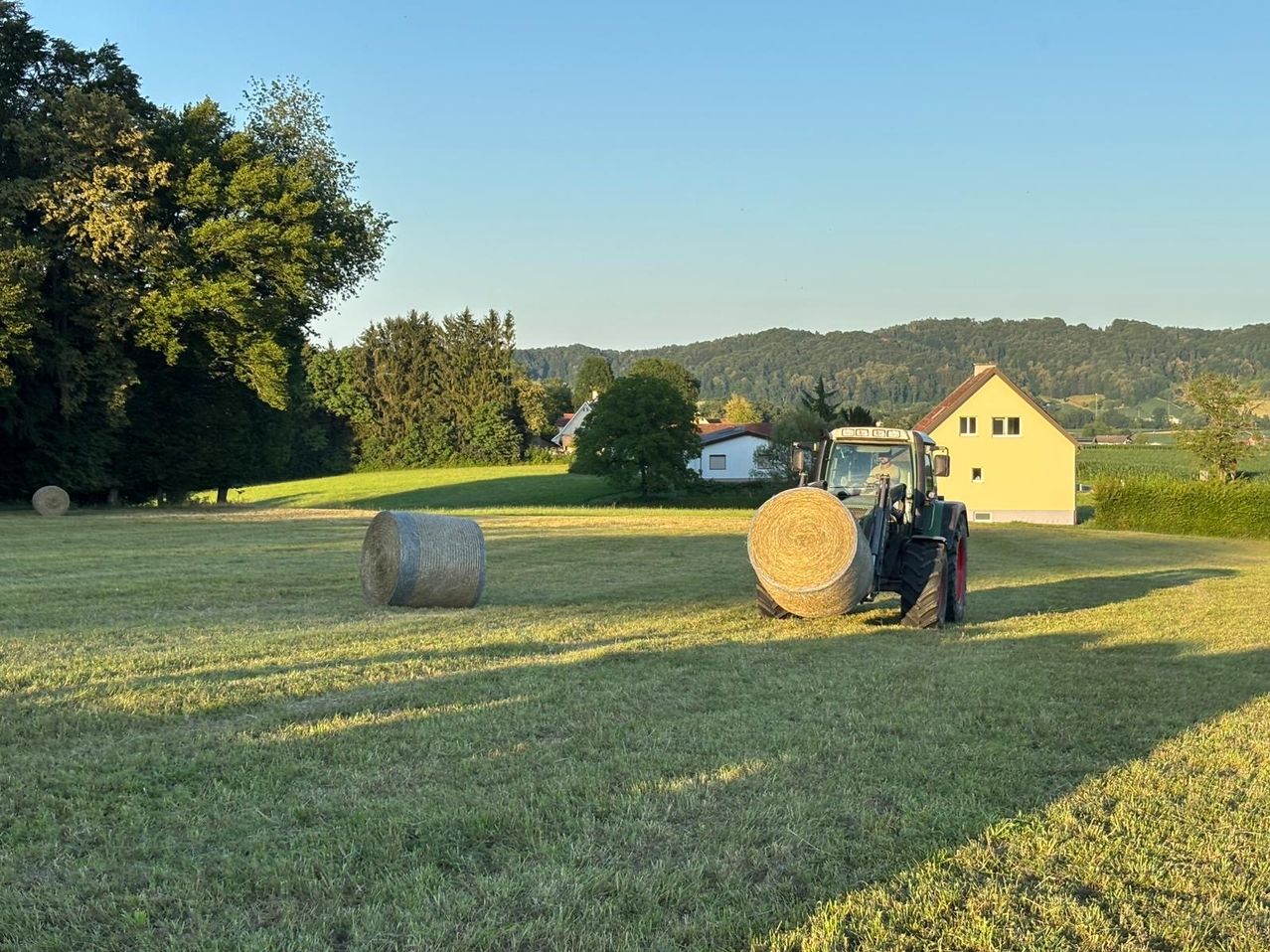 Ein Traktor erntet Heu auf einem Feld mit zwei großen Heuballen im Vordergrund. Ein gelbes Haus und ein weißes Haus sind im Hintergrund zu sehen, umgeben von Bäumen und Hügeln.