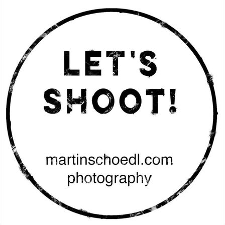 Ein runder Stempel in Schwarz und Weiß mit der Aufschrift 'Let's Shoot!'. Darunter befindet sich eine Website-Adresse, 'martinschoedl.com', und 'Fotografie' in kleinerer Schrift.