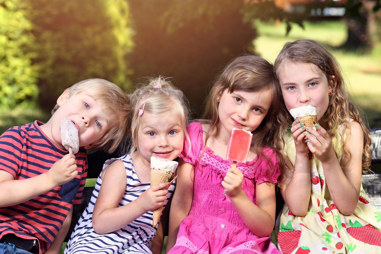Four children are sitting together and enjoying ice cream. Two are holding ice cream cones, and two are holding ice pops. They are all smiling and seem to be posing for a photo.