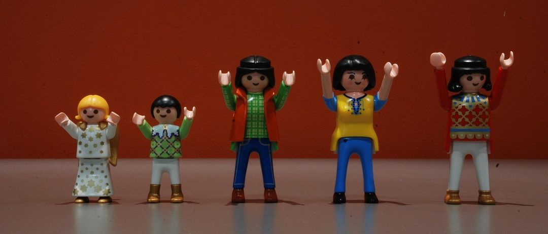 Fünf Playmobil-Figuren mit unterschiedlicher Kleidung und Posen stehen vor einem roten Hintergrund. Von links nach rechts sind die Figuren ein Mädchen in einem weißen Kleid, ein Junge in Grün, eine Frau in Rot, eine Frau in Blau und ein Mann in Rot.