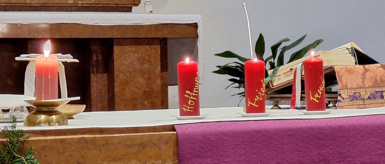 Ein Altar in einer Kirche mit vier brennenden roten Kerzen auf einem lila Tuch. Im Hintergrund befinden sich Blumen und Pflanzen. Die Kerzen tragen die Namen 'Hoffnung,' 'Friede,' 'Liebe' und 'Glaube'.