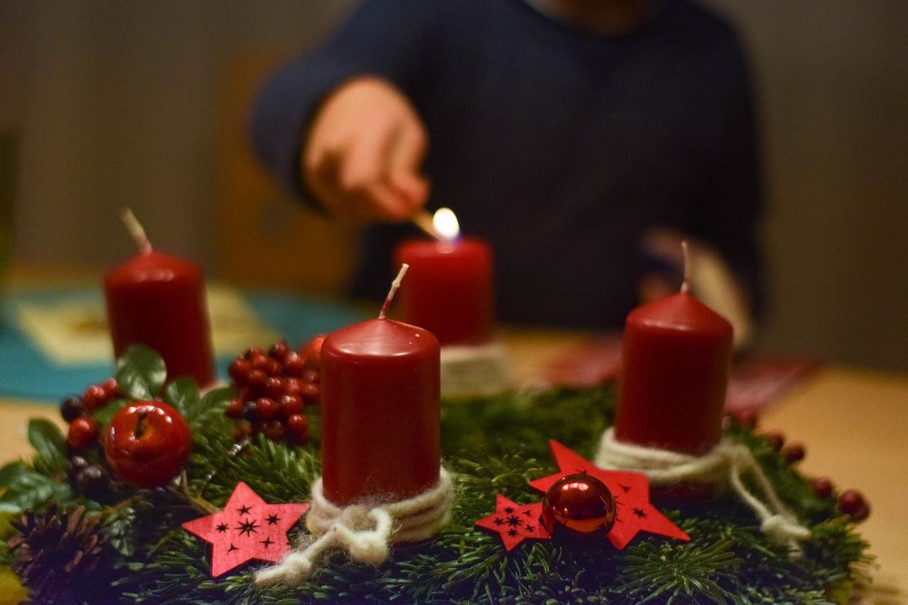 A child lights a red candle on a wreath with greenery, red stars, and small red balls. Four other red candles are partially lit on the wreath.