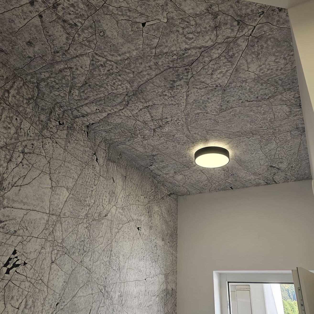 Ein Raum mit einer rissigen Musterdecke und einer weißen Wand. Eine runde Deckenleuchte ist montiert und eine weiße Tür ist teilweise geöffnet.