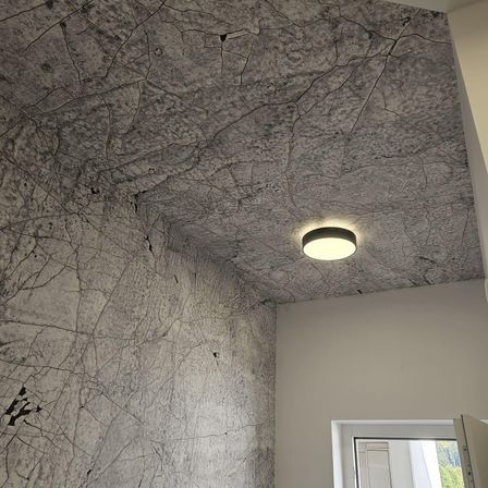 Ein Raum mit einer rissigen Musterdecke und einer weißen Wand. Eine runde Deckenleuchte ist montiert und eine weiße Tür ist teilweise geöffnet.