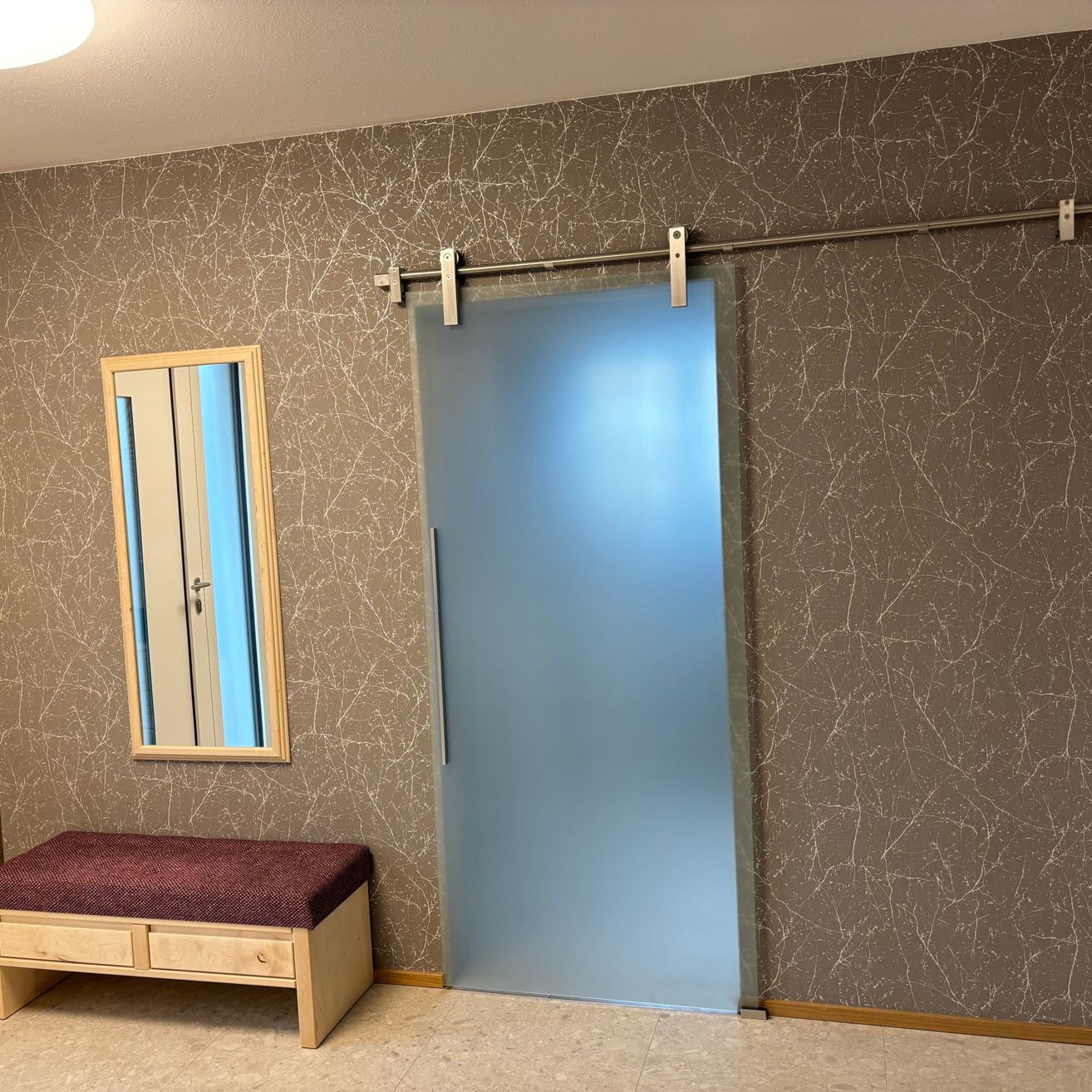 Ein leerer Raum mit einer Bank und einem Spiegel. Eine blaue Glastür befindet sich rechts, und dahinter ist eine hölzerne Wand mit Muster.