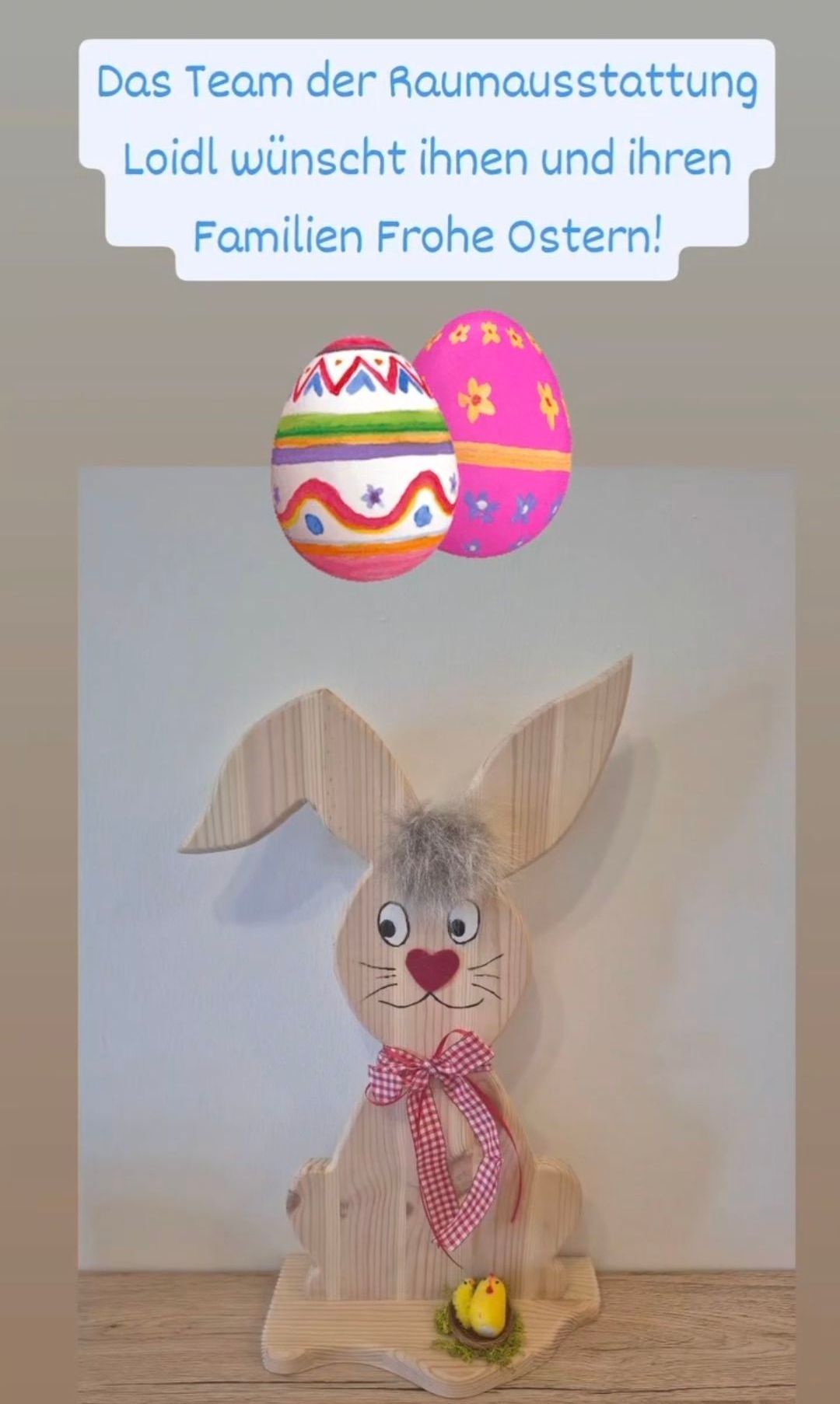 Ein hölzerner Osterhase mit rotem Band hängt an einer weißen Wand, darüber schweben zwei bunte Ostereier.