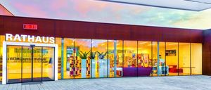 Das Äußere einer modernen Buchhandlung mit großen Glasfenstern, einer Glastür und Regalen voller Bücher. Der Eingang hat eine rote Wand mit dem Wort 'US'. Im Inneren gibt es ein Sofa und eine Ausstellung von Büchern.