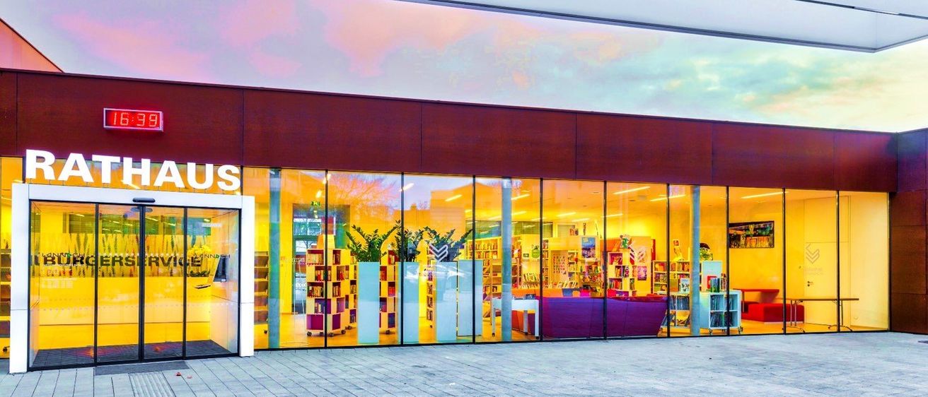 Ein Bibliotheksaußenbereich mit großen Glasfenstern, die Bücherregale, Pflanzen und einen Lesebereich mit einem lila Sofa zeigen. Der Bibliotheksname 'US' ist an der oberen Wand sichtbar.