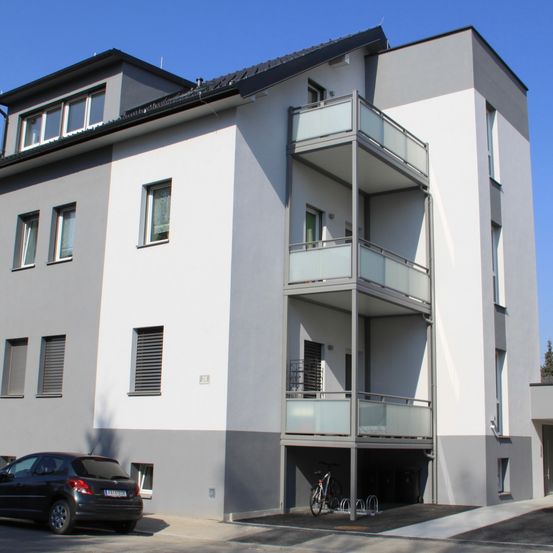 Ein modernes Mehrfamilienhaus mit weißen und grauen Außenwänden, mehreren Fenstern und Balkonen im zweiten Stock. Ein schwarzes Auto ist davor geparkt.