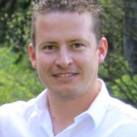 Nahaufnahme eines Mannes in einem weißen Hemd, der lächelnd in die Kamera schaut, mit einem Wald im Hintergrund.