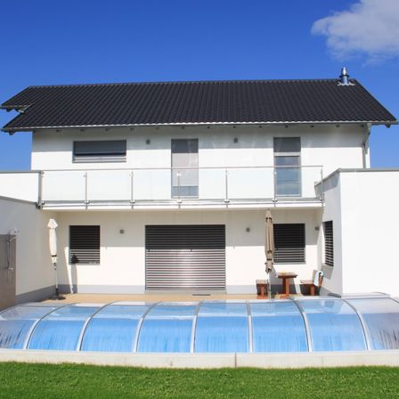 Ein modernes weißes Haus mit schwarzem Dach und einer Glaspoolabdeckung davor.