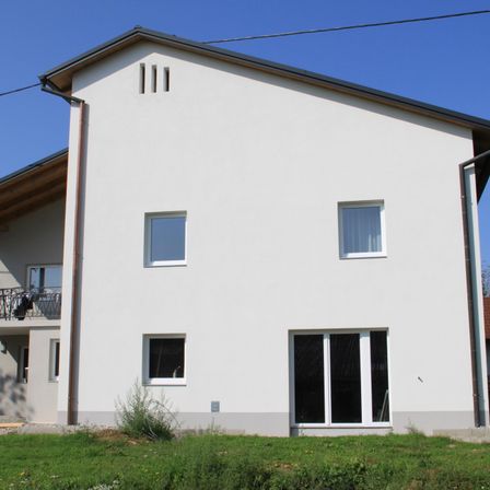 Ein zweistöckiges weißes Haus mit grauem Dach, Fenstern und einer Schiebetür im Erdgeschoss, einem Balkon mit Geländern im ersten Stock und einem Garten davor.
