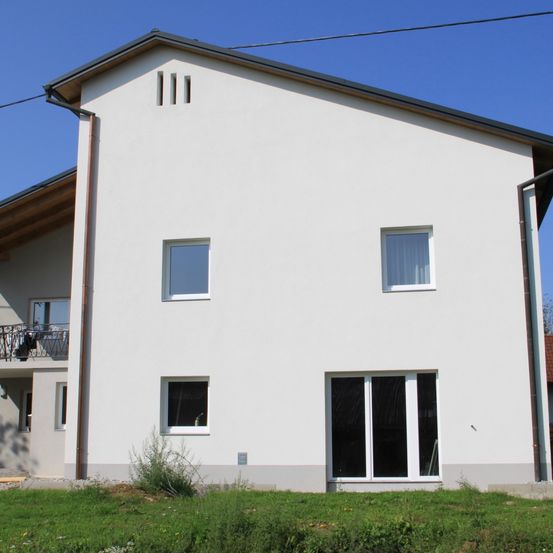 Ein zweistöckiges weißes Haus mit grauem Dach, Fenstern und einer Schiebetür im Erdgeschoss, einem Balkon mit Geländern im ersten Stock und einem Garten davor.