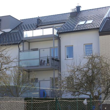 Ein modernes Haus mit einem gefliesten Dach und einem verglasten Balkon im zweiten Stock. Die Vorderseite des Hauses hat einen Schornstein und einen Zaun.