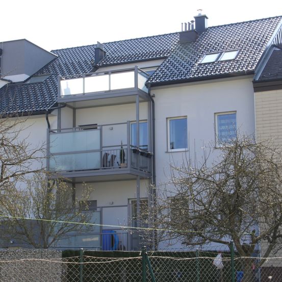 Ein modernes Haus mit einem gefliesten Dach und einem verglasten Balkon im zweiten Stock. Die Vorderseite des Hauses hat einen Schornstein und einen Zaun.