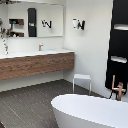 Ein modernes Badezimmer mit einer weißen Badewanne, einem Holzschrank mit Waschbecken, einem Spiegel und einem Kupferhahn. Der Boden ist mit grauen Fliesen belegt.