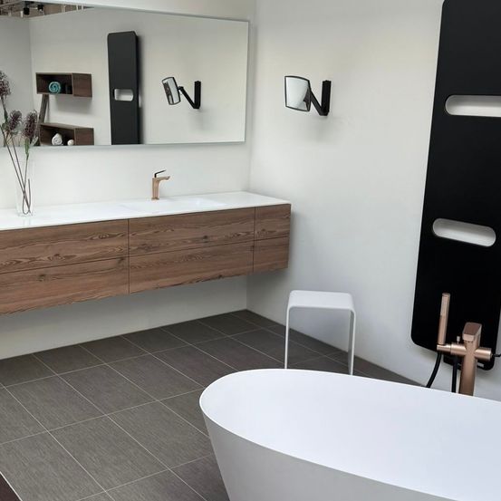 Ein modernes Badezimmer mit einer weißen Badewanne, einem Holzschrank mit Waschbecken, einem Spiegel und einem Kupferhahn. Der Boden ist mit grauen Fliesen belegt.