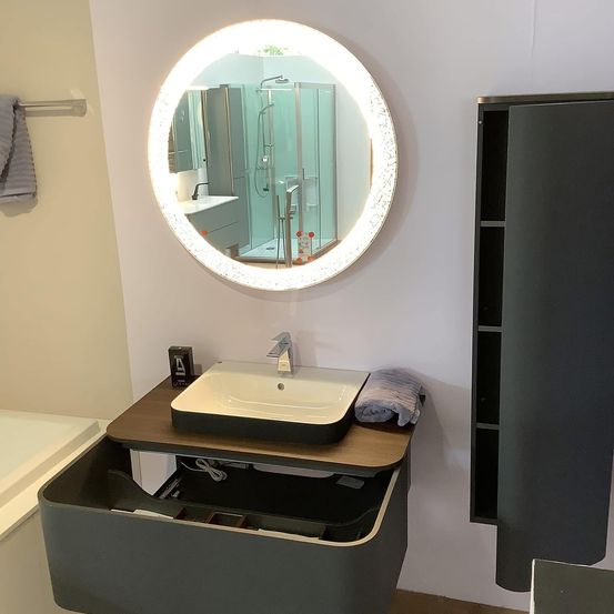 Ein Badezimmer mit einem großen beleuchteten Spiegel, einem Waschbecken, einem Handtuchhalter und einem Schrank. Der Spiegel reflektiert den Duschbereich.