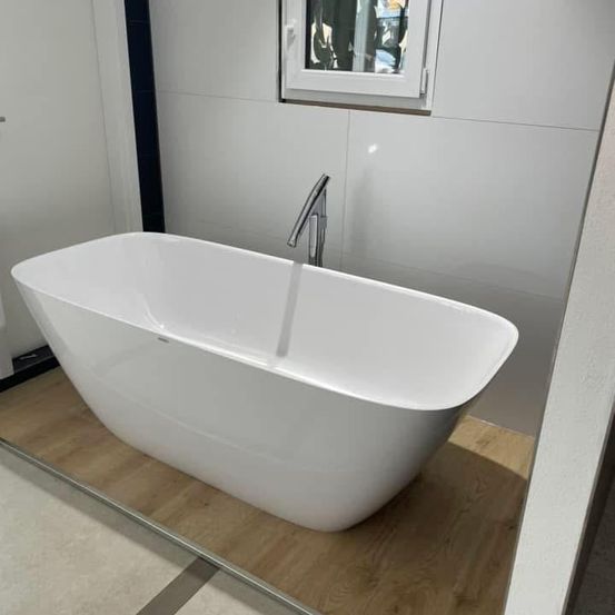 Eine weiße Badewanne mit einem Wasserhahn steht auf einem Holzboden in einem Badezimmer mit weißen Wänden und einem Fenster.