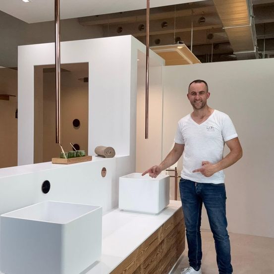 Ein Mann in einem weißen T-Shirt und Jeans steht in einem Badezimmer und zeigt auf ein weißes Waschbecken.