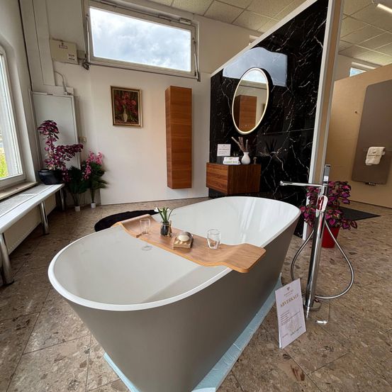 Ein modernes Badezimmer mit einer großen weißen Badewanne mit einem Holztablett, Gläsern und einer Seife. Der Raum hat eine Marmorwand, einen Spiegel und einen Holzschrank mit einer Vase. Es gibt Topfpflanzen und eine Bank am Fenster.