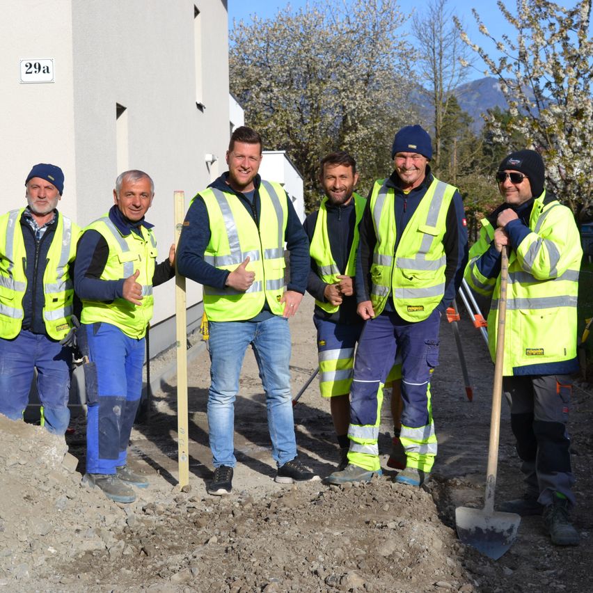 Fünf Männer in Sicherheitswesten und Schutzhelmen stehen auf einem unbefestigten Weg und posieren für ein Foto. Ein Mann hält eine Schaufel und ein anderer einen Holzpfahl. Hinter ihnen steht ein Gebäude mit der Nummer 29a an der Wand und einem Berg im Hintergrund.