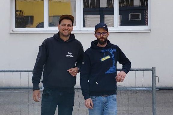 Zwei Männer in Kapuzenpullovern stehen nebeneinander vor einem weißen Gebäude. Einer hat ein lächelndes Gesicht und der andere trägt eine Brille.