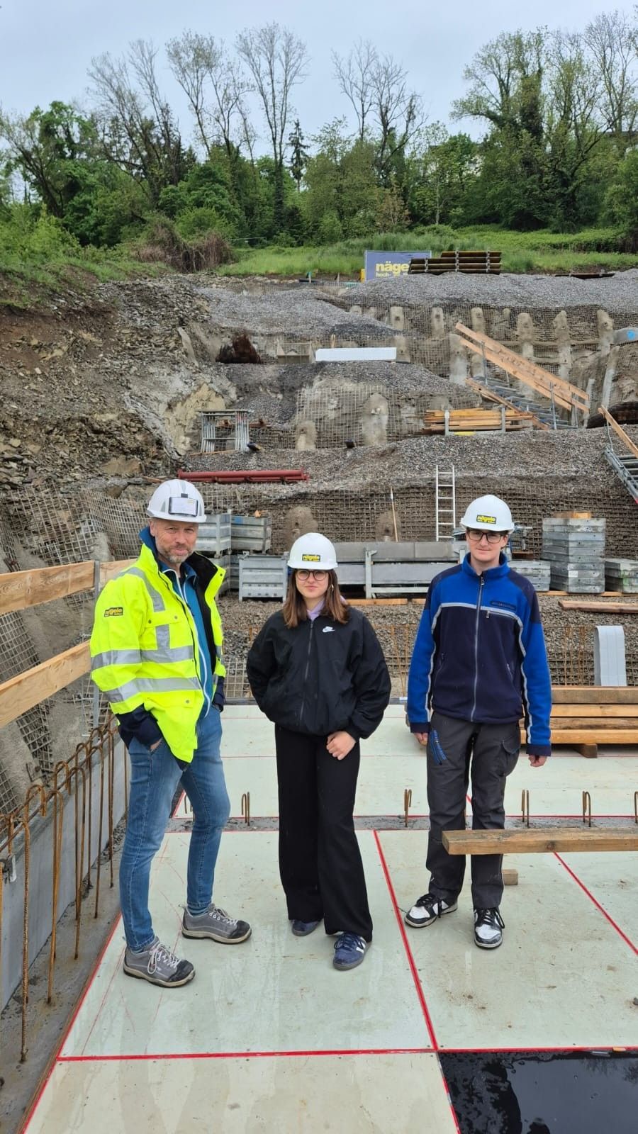 Drei Personen in Schutzhelmen und Arbeitskleidung posieren für ein Foto an einer Baustelle. Hinter ihnen sind ein Haufen Kies und Stahlstangen zu sehen, und eine Leiter lehnt an einer Wand.