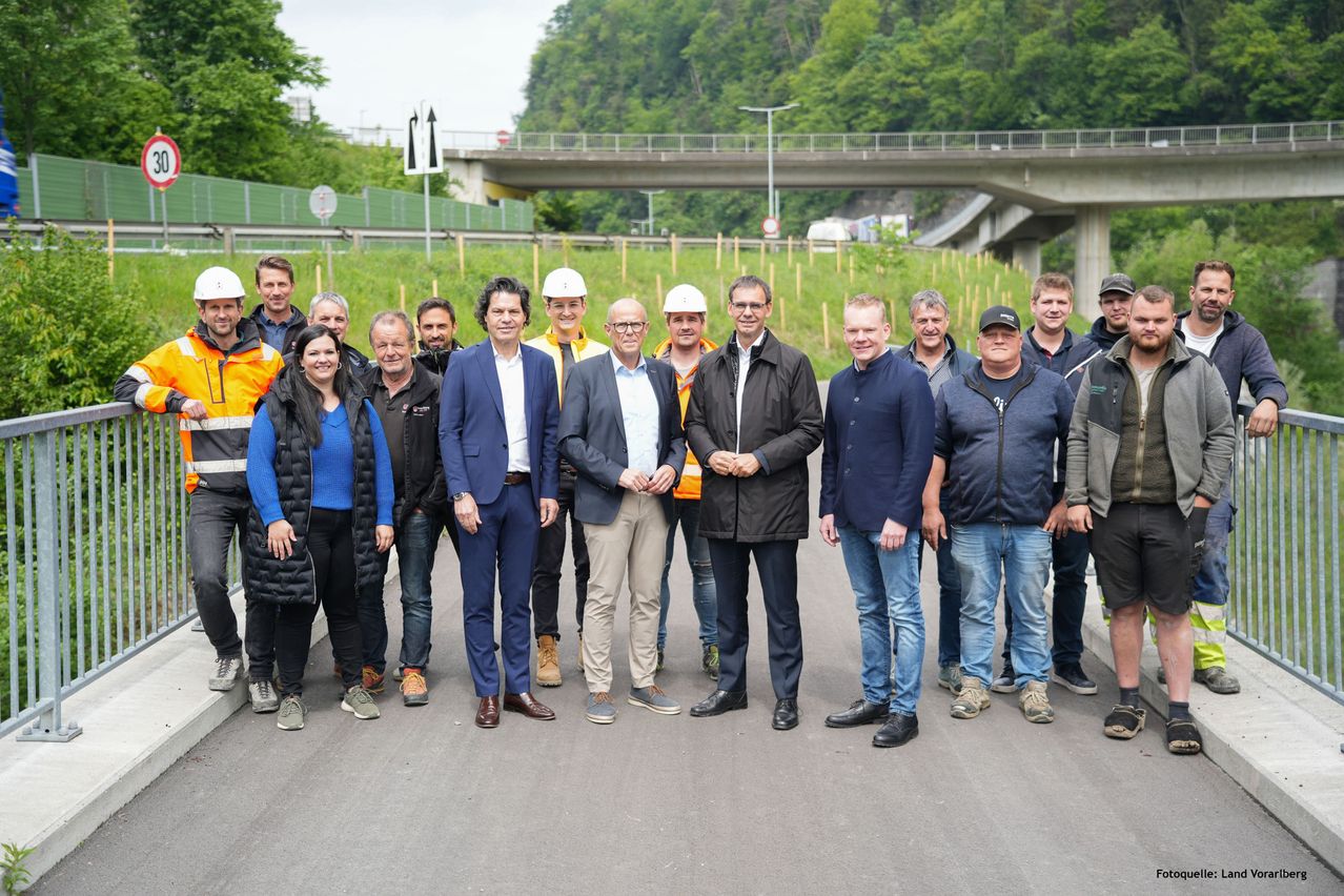 Eine Gruppe von Menschen in Jacken und Mützen steht vor einer Baustelle, mit einer Brücke im Hintergrund.