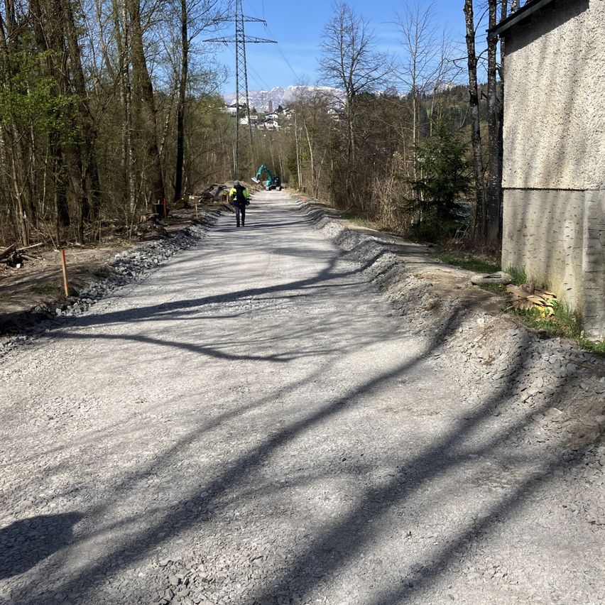 Eine Person geht auf einem Schotterweg, gesäumt von Bäumen. In der Ferne ist ein Baufahrzeug zu sehen. Im Hintergrund sind Berge zu erkennen.