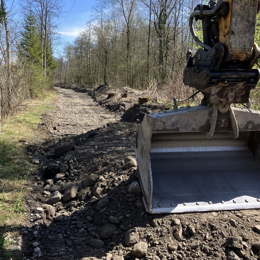 Eine Baustelle mit einem großen Bagger, der an einem von Bäumen umgebenen Erdweg arbeitet.