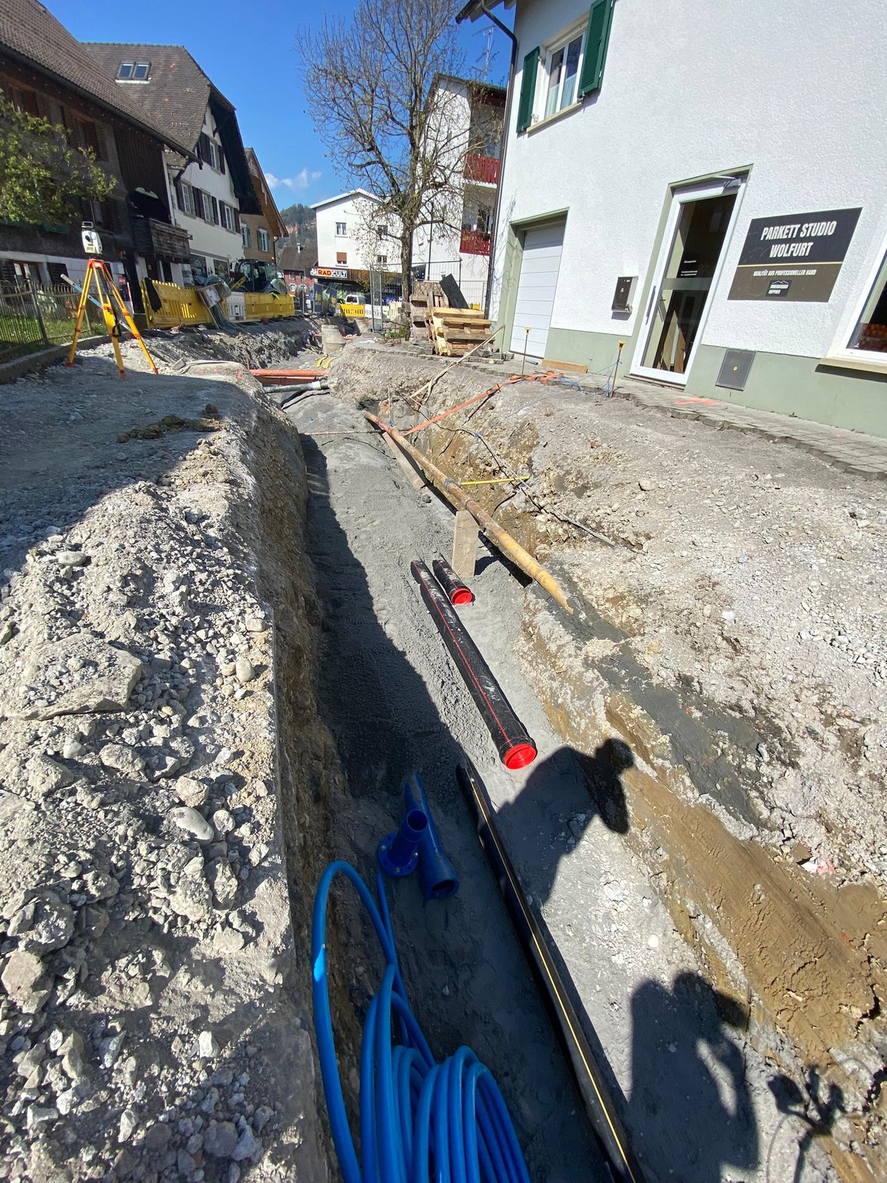 Baustelle mit einem Graben in einem Dorf, auf dem eine neue Rohrleitungsinstallation zu sehen ist. Ein Gebäude mit dem Schild 'Parrett Studio Wolfurt' ist sichtbar.