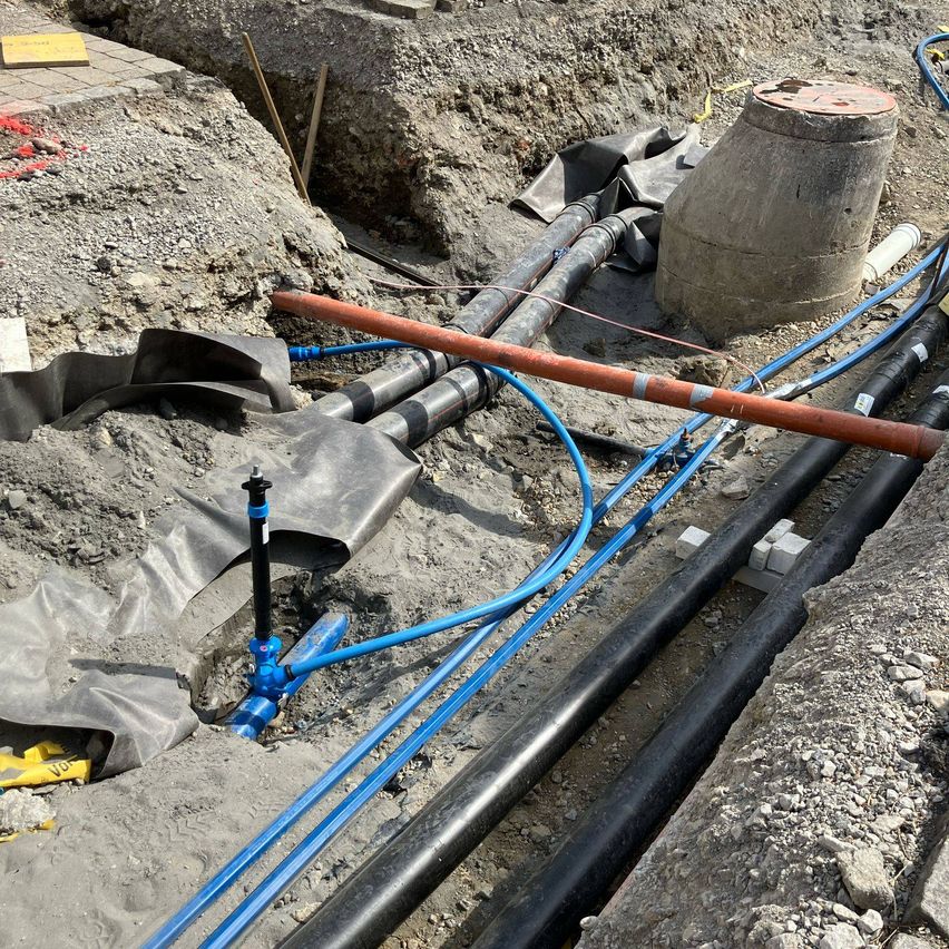 Ein Baustelle mit freiliegenden Rohren und einem Betontank. Mehrere blaue und schwarze Rohre liegen auf dem Boden. Ein Holzstab ist links sichtbar.
