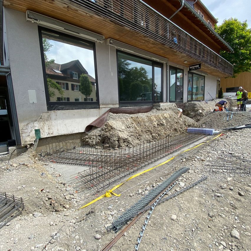 Eine Baustelle vor einem Gebäude mit reflektierenden Fenstern. Arbeiter sind auf der Baustelle tätig, einer gräbt und der andere steht. Es gibt Haufen von Erde, Baumaterialien und gelbes Warnband.