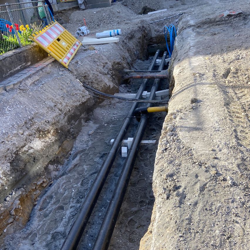 Eine Baustelle im Freien mit einem ausgehobenen Graben für die Verlegung von Rohrleitungen. Zwei große schwarze Rohre liegen mit weißen Stützen. Ein gelbes Schild befindet sich links.