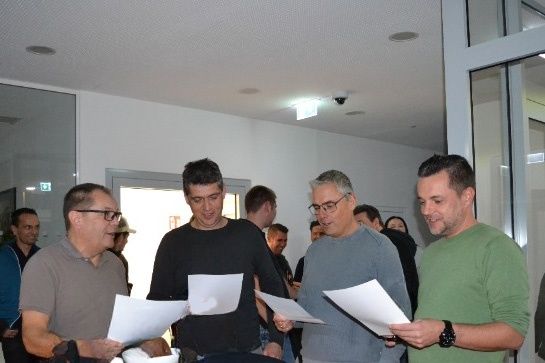 Eine Gruppe von Männern ist in einem Raum versammelt, hält Papiere in der Hand und scheint etwas zu besprechen. Ein Mann trägt eine Brille und eine Uhr.