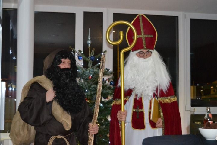 Zwei Personen in Kostümen stehen vor einem Weihnachtsbaum. Einer hält einen Stab und ein Buch, der andere einen Stock und eine Tasche. Hinter ihnen befindet sich ein Fenster.