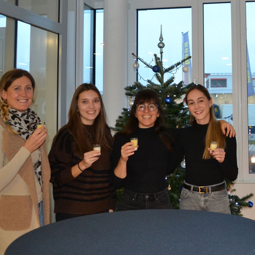 Vier Frauen stehen lächelnd vor einem geschmückten Weihnachtsbaum und halten kleine Tassen. Sie befinden sich in einem Büro mit großen Fenstern und einem blauen Tisch davor.
