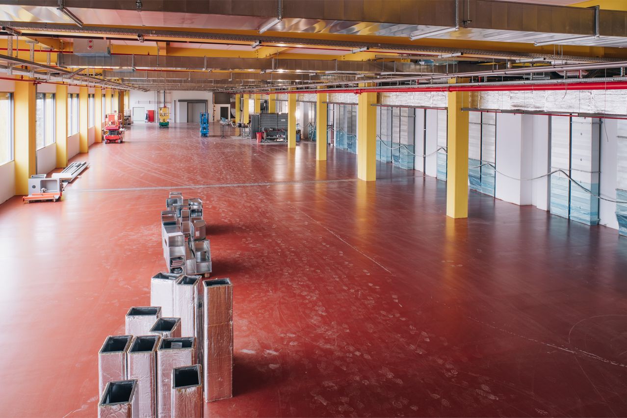 Ein leerer Fabrikboden mit roter Farbe, Metallgegenständen und gelben Säulen. Die Decke hat Lichter und Maschinen.