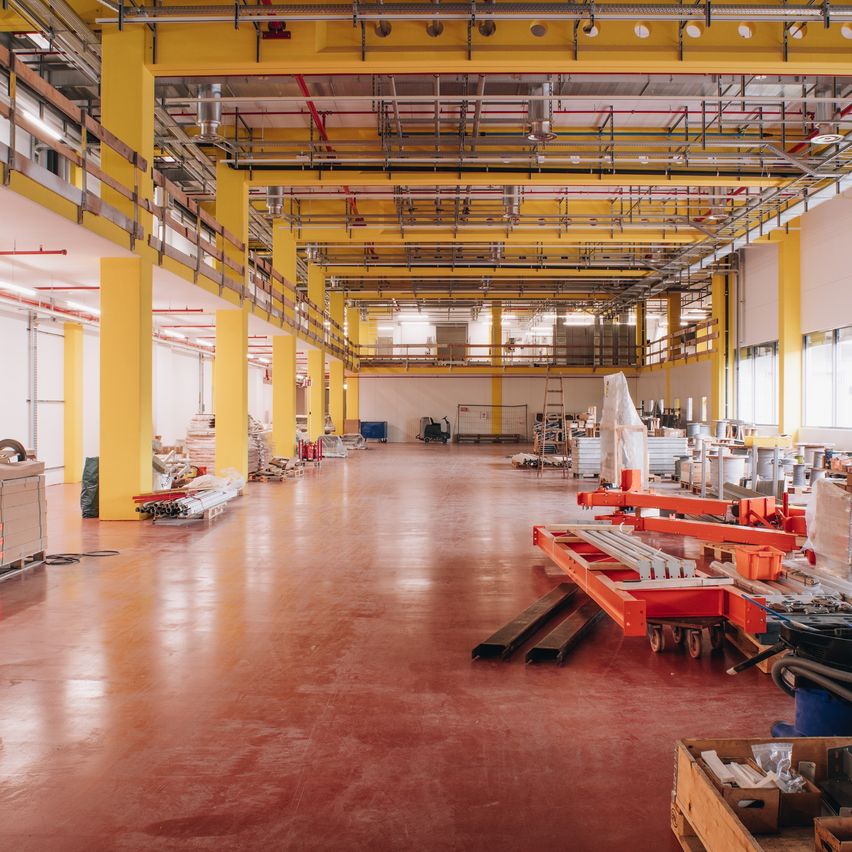 Ein leeres Lagerhaus mit gelben Säulen und rotem Boden. Die Decke hat Metallträger und Beleuchtung. Es gibt mehrere Gegenstände und Maschinen im Bereich.