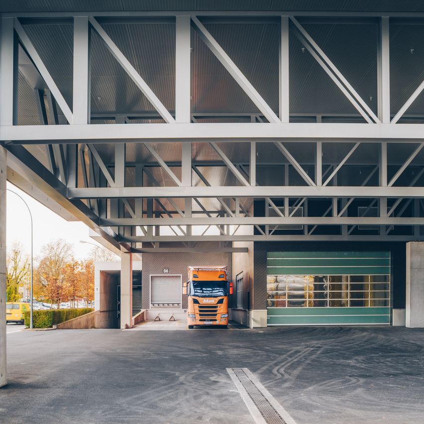 Ein leerer Parkplatz mit einem geparkten LKW. Der Bereich befindet sich unter einer Metallstruktur mit Balken und einem Dach. Bäume und Fahrzeuge sind in der Ferne.