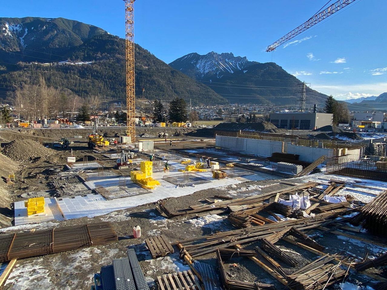 A construction site with cranes, workers, yellow barriers, wooden planks, and piles of dirt. Mountains with snow in the background.