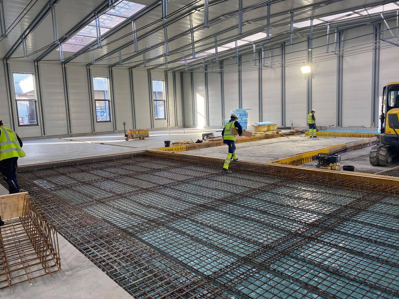 Ein Innenbaustelle mit Arbeitern in Sicherheitsausrüstung. Stahlarmierungen liegen auf dem Boden. Verschiedene Werkzeuge und Materialien sind verstreut.