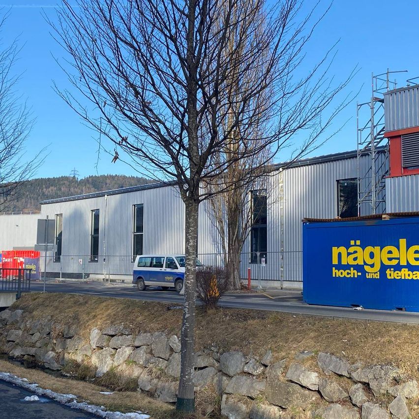 Ein blaues Gebäude mit Nagele Hoch- und Tiefbau Schild steht neben einer Steinmauer und einem kahlen Baum. Ein blauer und weißer Van ist vor dem Gebäude geparkt.