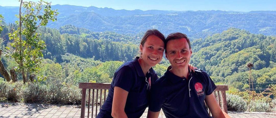 Zwei Personen sitzen unter roten Regenschirmen am Tisch und lächeln. Der Tisch ist mit Tellern, Gläsern und Gläsern gedeckt. Hinter ihnen sind Berge und Grünflächen sichtbar.