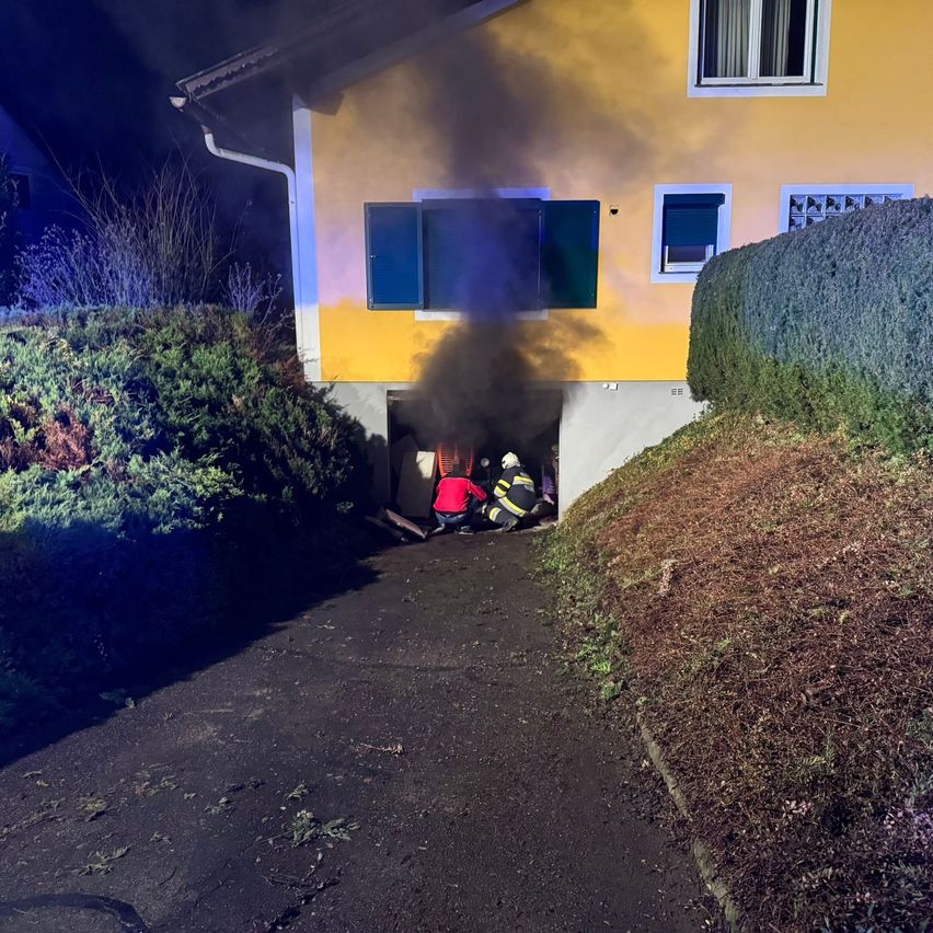 Zwei Feuerwehrleute stehen vor einem gelben Haus, aus dessen Garagentür Rauch aufsteigt. Eine Person sitzt neben ihnen auf dem Boden.