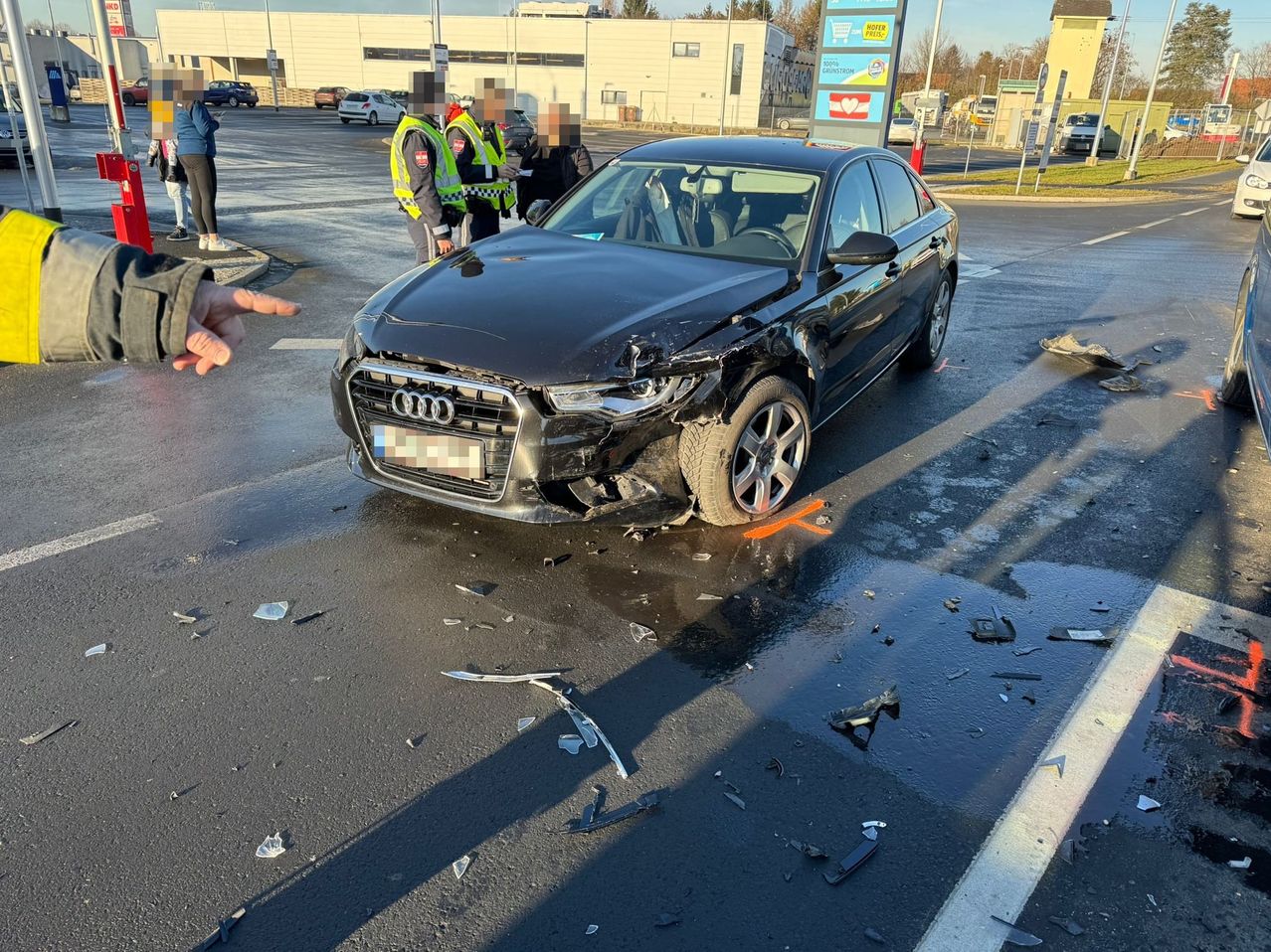 Ein schwarzer Audi-Limousine ist auf der Straße demoliert, mit zerbrochenem Glas und Trümmern, wobei zwei Personen in Sicherheitswesten in der Nähe stehen.