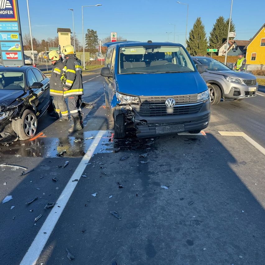 Ein blauer Volkswagen-Transporter ist vorne auf der Straße beschädigt. Zwei Feuerwehrleute stehen in der Nähe. Ein schwarzes Auto ist links demoliert und ein silbernes Auto ist rechts geparkt.