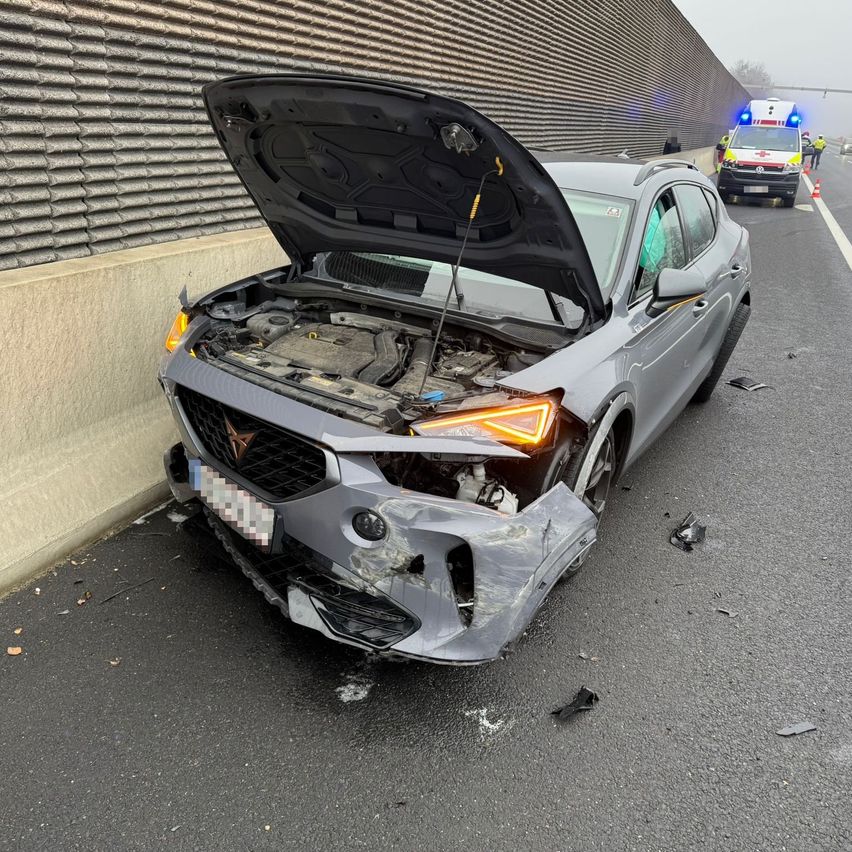 Ein grauer SUV steht mit offenem Motorhauben an der Straßenseite. Der vordere Stoßfänger ist beschädigt. Dahinter ist ein Rettungswagen geparkt.
