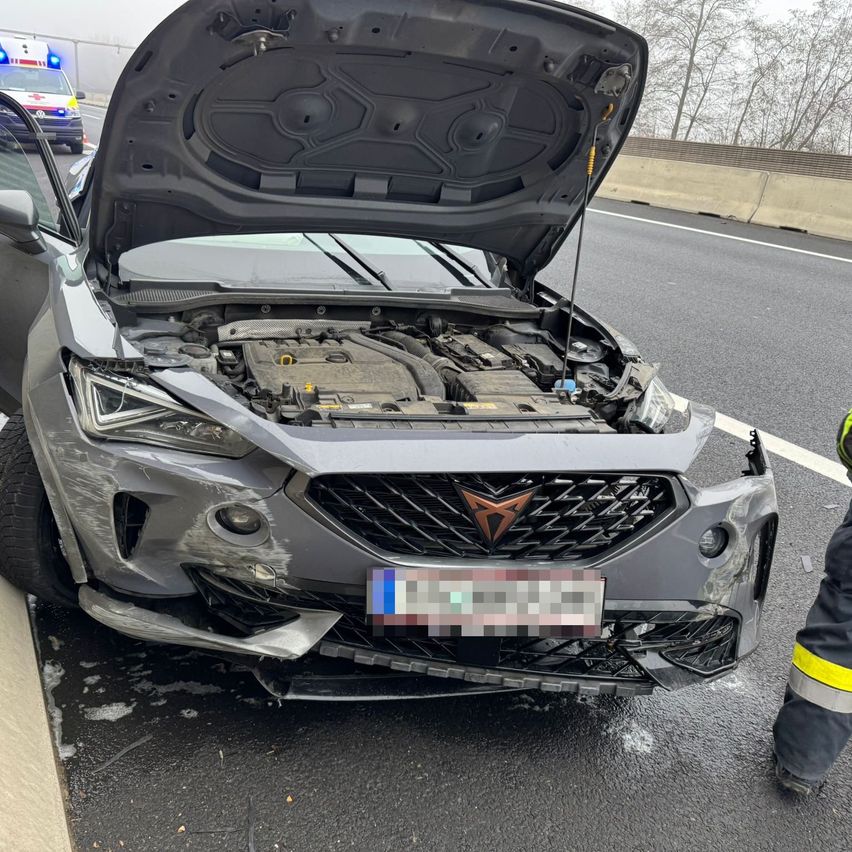 Ein graues Auto mit offenem Motorhaube steht am Straßenrand, mit sichtbaren Schäden an der vorderen Stoßstange. Ein Rettungsfahrzeug ist im Hintergrund zu sehen.